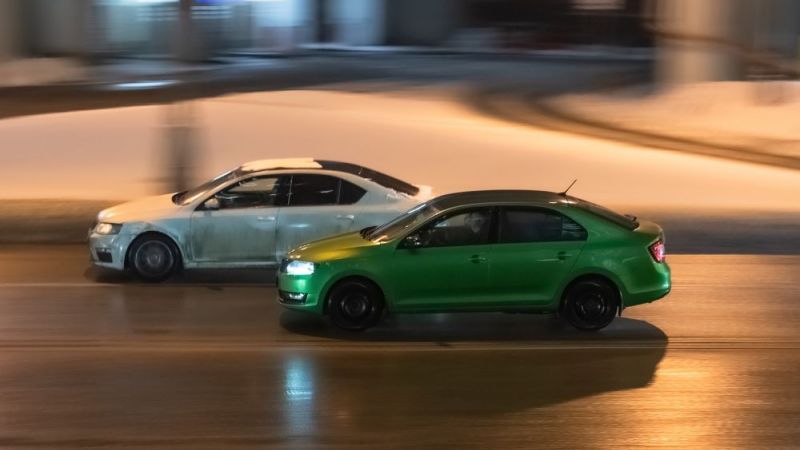 Two cars in motion on city street at night. Racing at high speed on public road. White and green sedans drive on wet winter road