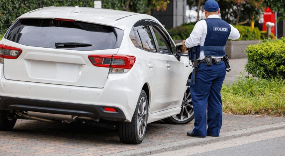 A white car is parked on a cobblestone street. A police officer stands beside it, inspecting, in a leafy residential area. The officer's vest reads "POLICE."