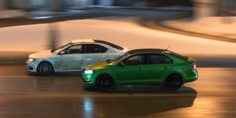 Two cars in motion on city street at night. Racing at high speed on public road. White and green sedans drive on wet winter road