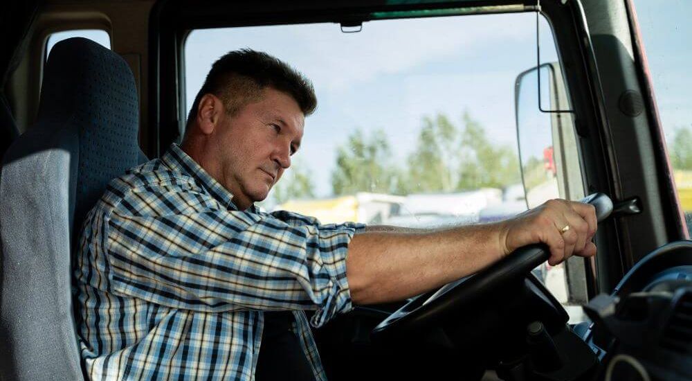 A person in a plaid shirt drives a vehicle, focusing intently. The interior is dimly lit with a window showing a blurred, sunny outdoor scene with trees.