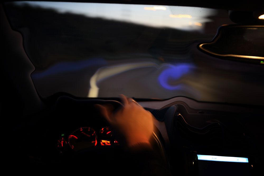 A driver's hand on a steering wheel inside a car at night, illuminated dashboard visible, blurring scenery outside indicates motion, with the road curving to the left.