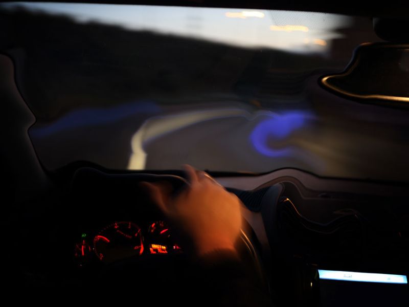 A driver's hand on a steering wheel inside a car at night, illuminated dashboard visible, blurring scenery outside indicates motion, with the road curving to the left.