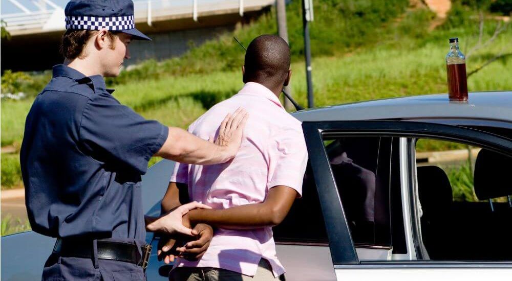 Police officer handcuffing driver during roadside DUI arrest next to car with a bottle of alcohol on the roof.