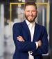 Calum Welch, criminal defense attorney with folded arms wearing a blue suit and white shirt stands confidently, framed by a golden outline, against a blurred office background.