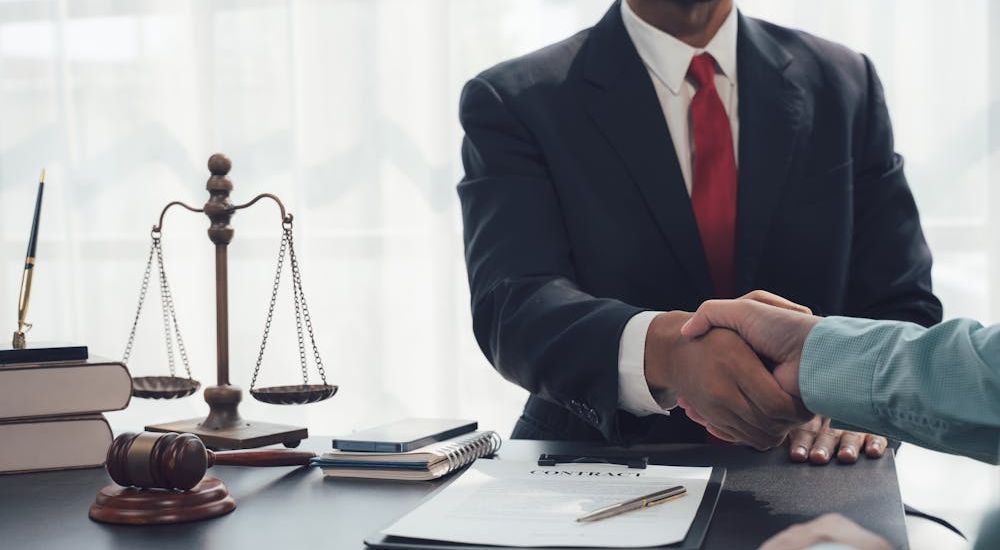 Two people shaking hands across a desk with law-related items like scales, a gavel, books, and a document labeled "CONTRACT" in an office setting.