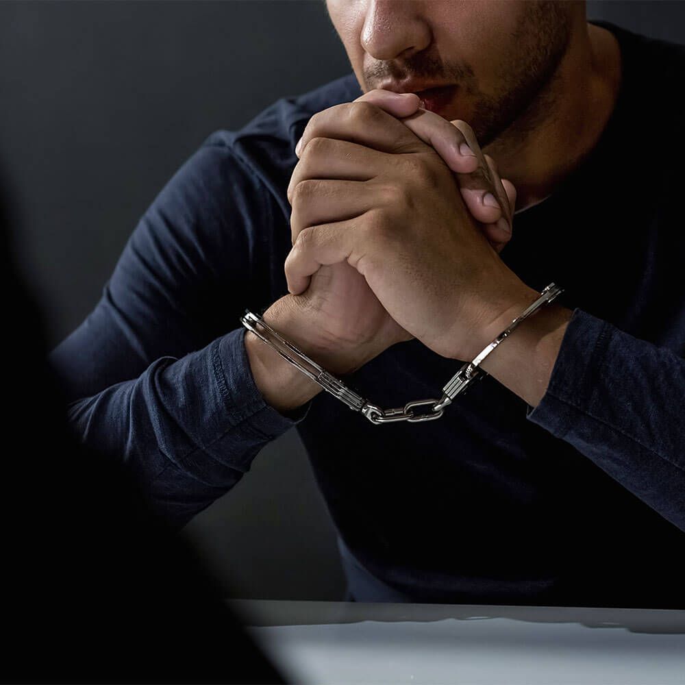 Handcuffed man seated at a table with hands clasped, appearing tense and contemplative during questioning.
