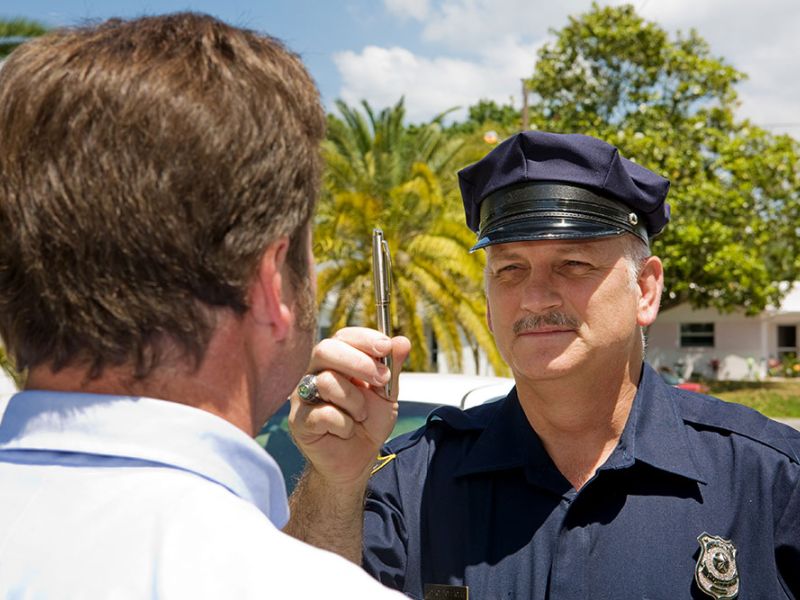 A police officer is performing a sobriety test on a man by holding out a pen, against a suburban backdrop with palm trees.