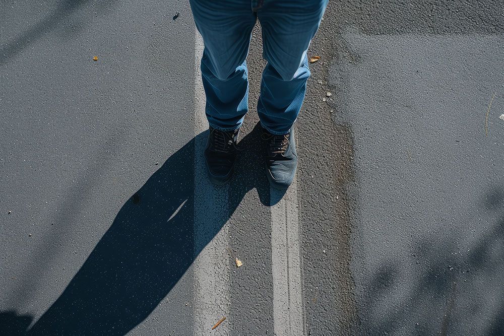 A person stands on an asphalt surface with a strong shadow, denoting sunlit conditions, possibly signifying a pause or contemplation.