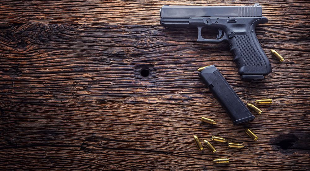 A handgun lies adjacent to a detached magazine and scattered ammunition on a textured wooden surface.