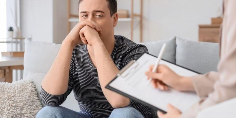 Anxious man sits on a couch with hands clasped, while a therapist takes notes on a clipboard nearby.