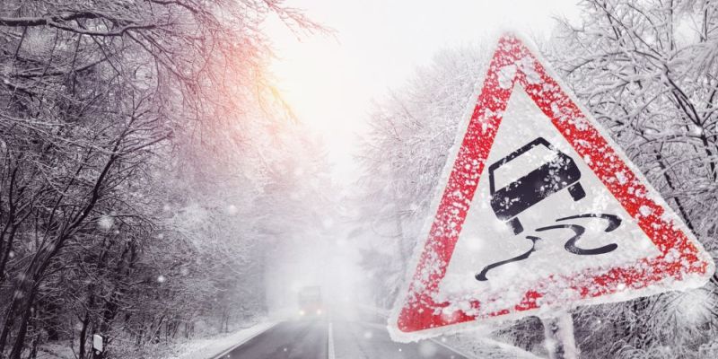 Winter forest road blanketed in snow with a triangular slippery-road warning sign alerting drivers to icy conditions.