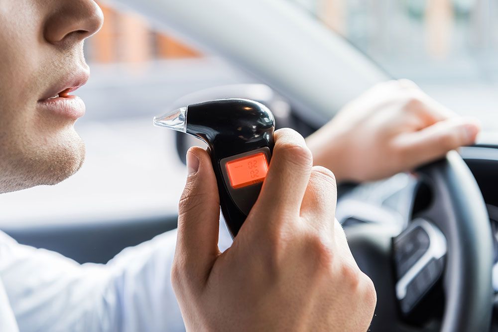 A man is using a breathalyzer in a car, his hand on the wheel, the device reads "0.02 BAC."