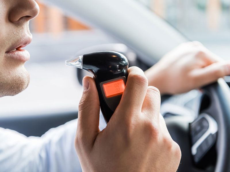 A man is using a breathalyzer in a car, his hand on the wheel, the device reads "0.02 BAC."