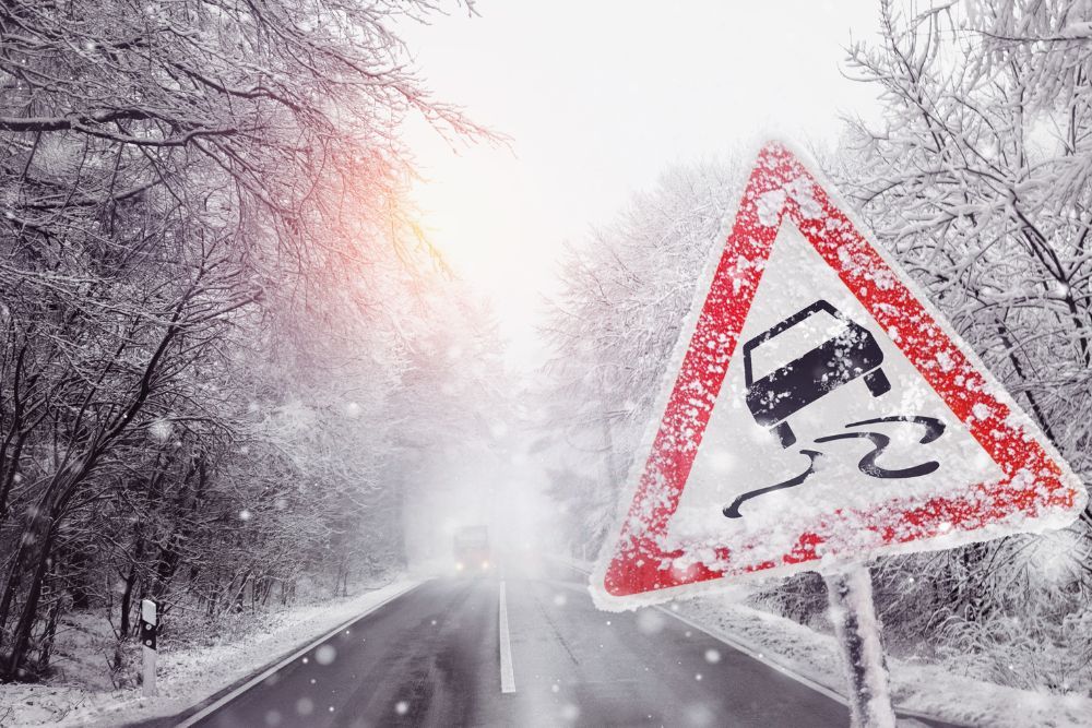 Winter forest road blanketed in snow with a triangular slippery-road warning sign alerting drivers to icy conditions.