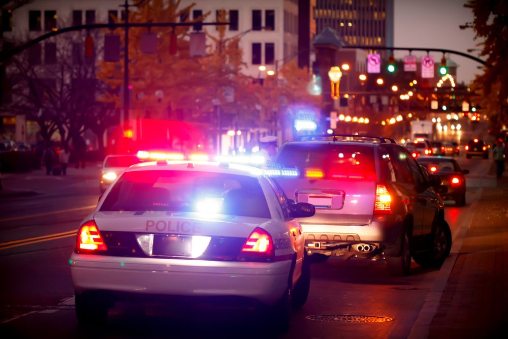 A police car with flashing lights stops behind a vehicle on a city street at dusk, surrounded by buildings and streetlights.