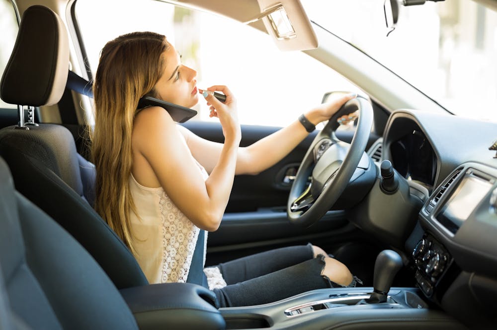 A woman sits in a car, driving with one hand on the wheel while holding a phone to her ear with the other. The interior is modern and clean, emphasizing multitasking.