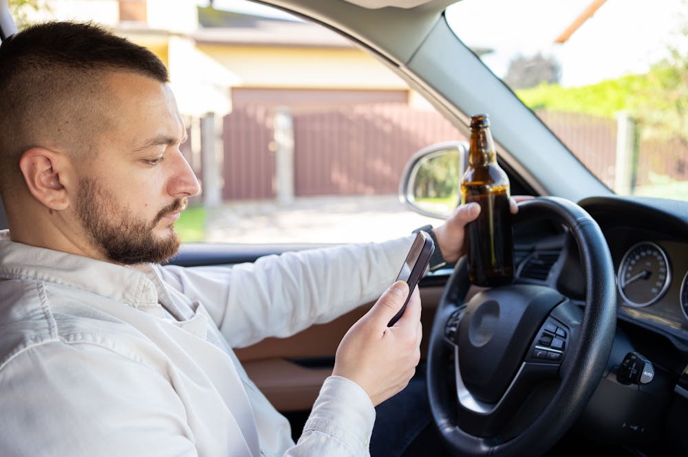 A man sits in a parked car holding a smartphone and a beer bottle, appearing engaged with the phone. The background features a gated residential area.