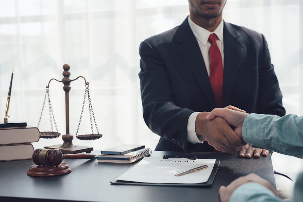 Two people shaking hands across a desk with law-related items like scales, a gavel, books, and a document labeled "CONTRACT" in an office setting.