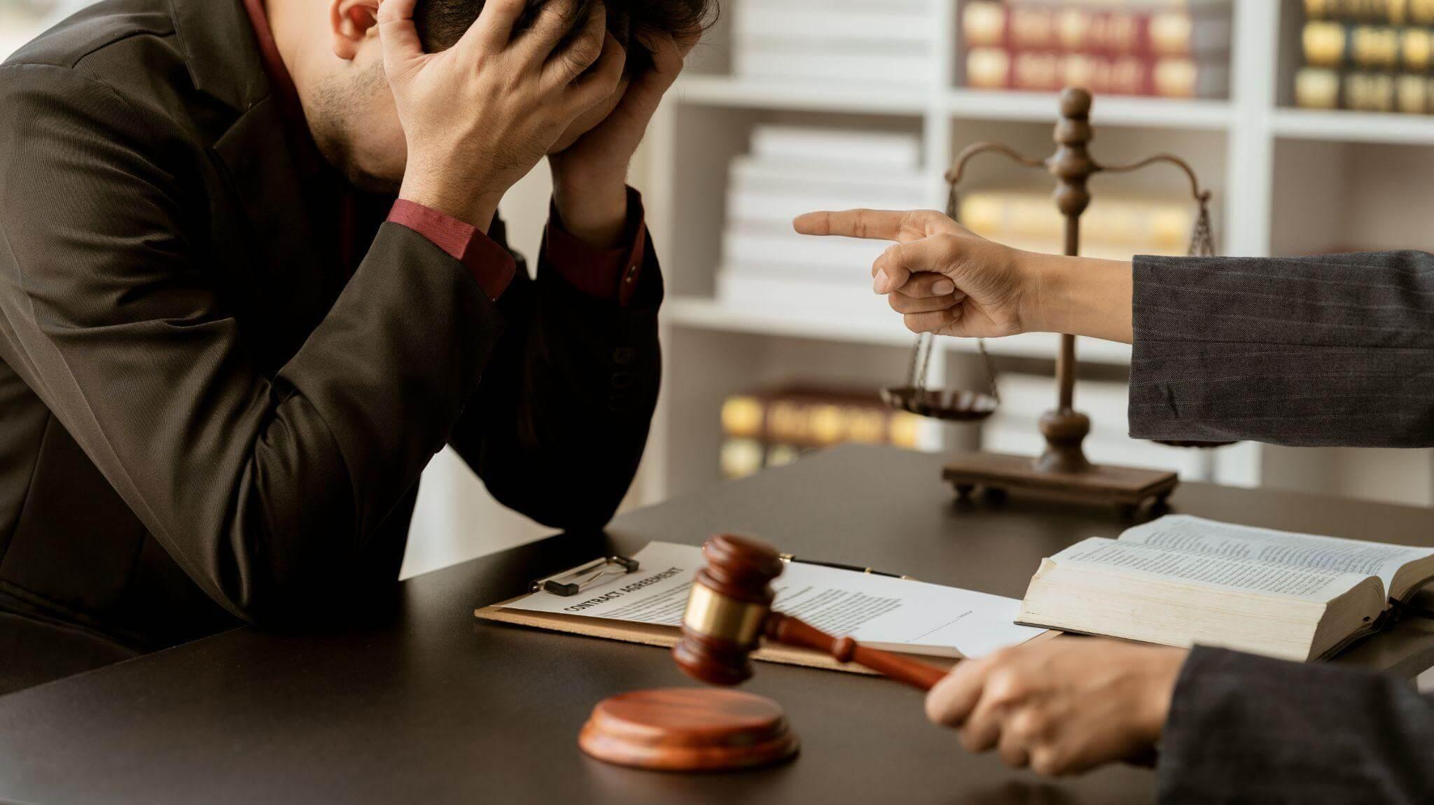 Man sitting at table in courtroom setting while a finger is pointing at him