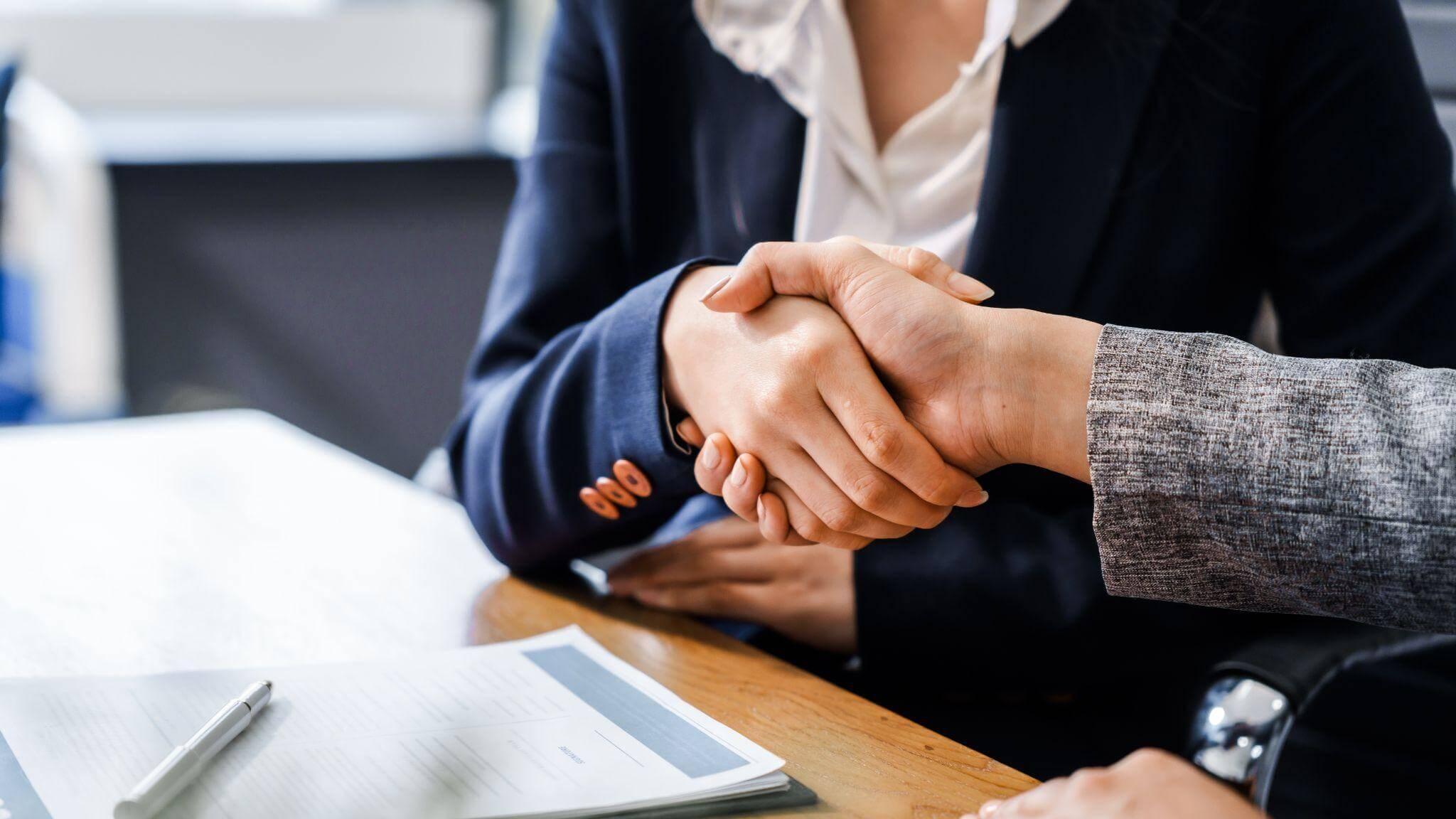 two people sitting at a table shaking hands, there's a document on the table and a pen on top of it